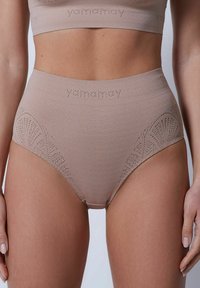 Högmidjad underkläder i mjuk beige tyg har ett strukturerat mönster och utskärningar på sidorna, med varumärkesnamnet diskret synligt.