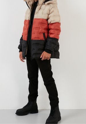 Lapsi, joka pukeutuu kolmiväriseen toppatakkiin, jossa on beige, punainen ja musta osio, mustat housut ja mustat nauhalliset saappaat, seisoo valkoista taustaa vasten.