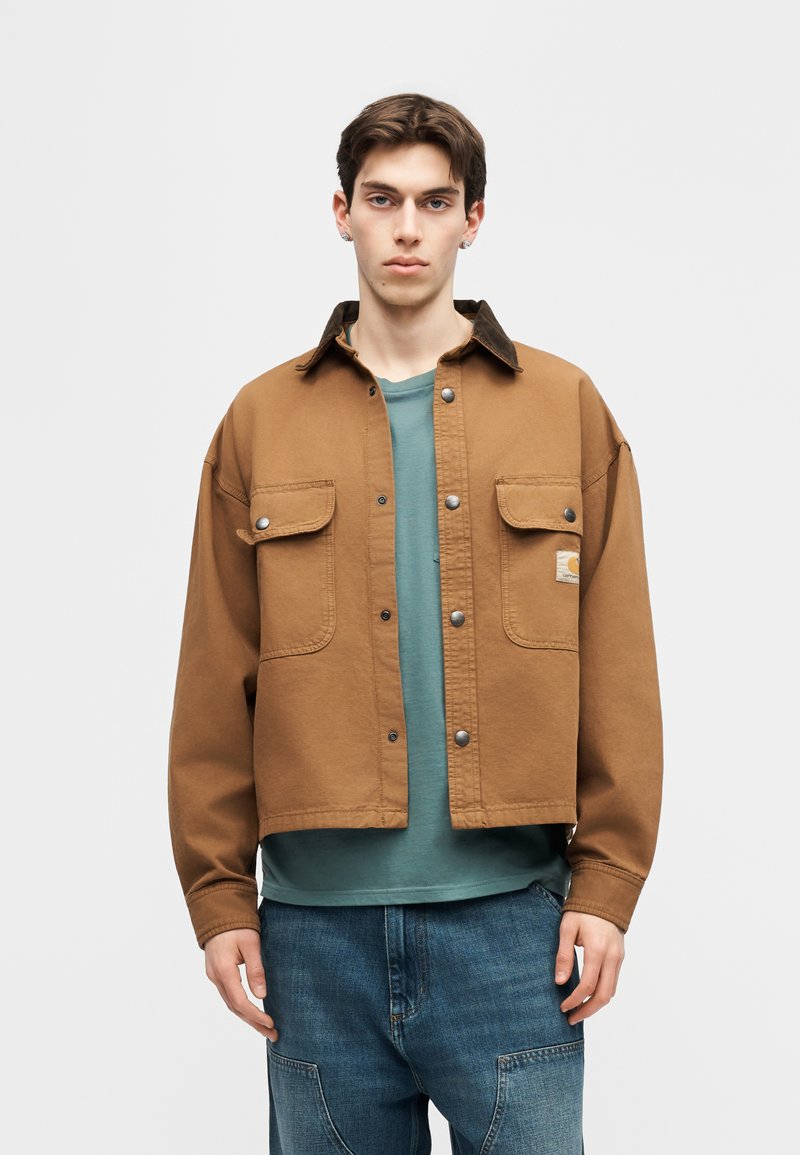 Junger Mann mit kurzen braunen Haaren trägt eine braune Jacke mit Knöpfen über einem türkisfarbenen Hemd und blauer Jeans, steht vor grauem Hintergrund.