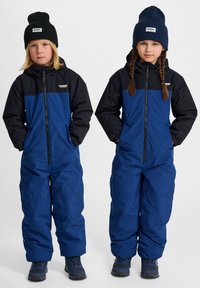 Monos integrales aislantes en azul y negro con frentes con cremallera, puños elásticos y bolsillos laterales. Gorras de punto en verde oscuro y azul marino.