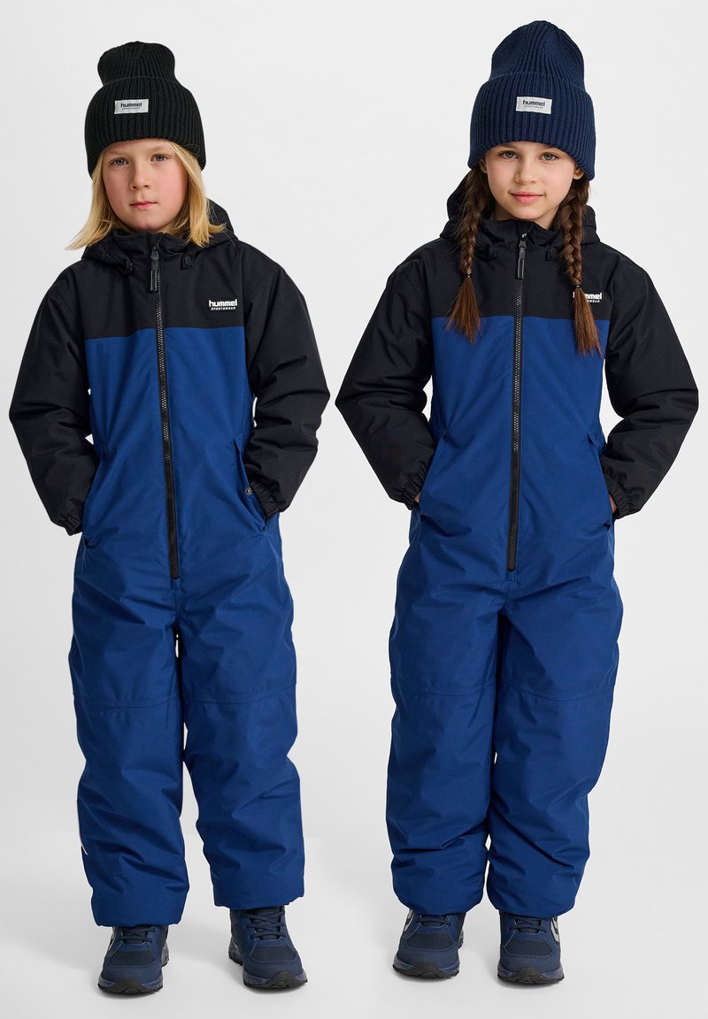 Monos integrales aislantes en azul y negro con frentes con cremallera, puños elásticos y bolsillos laterales. Gorras de punto en verde oscuro y azul marino.