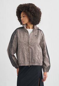 Καφέ μπουφάν με σχέδιο ζώου, με φερμουάρ μπροστά, ψηλό γιακά και ελαστικά μανσέτια. Μαύρες ρίγες κατά μήκος των μανικιών. Διαθέτει χρυσό φερμουάρ.