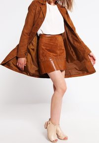 Brun mocka trenchcoat, vit topp och en brun minikjol med glansiga accenter och nitar. Modellen bär beigefärgade ankelskor.
