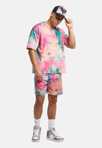 Farverig oversized skjorte og matchende shorts i pink og blå mønstre, parret med en caps. Skoene har livlige designs; fremtrædende logoer er synlige.