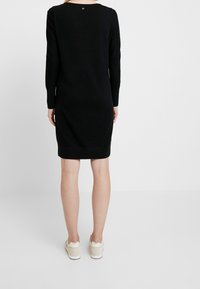 Robe noire en maille à manches longues avec un col rond, dotée d'un ourlet côtelé et d'un détail subtil de logo au dos. Portée avec des baskets claires.