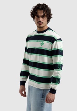 Man met krullend haar, gekleed in een wit, marineblauw en groen gestreept shirt met lange mouwen en tekst op de mouw, gecombineerd met lichtblauwe spijkerbroek.