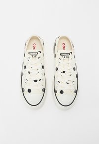 Białe tenisówki z czarnymi kropkami, zaokrąglonym noskiem, czarną gumową podeszwą i białymi sznurowadłami. Logo marki Converse na języku.