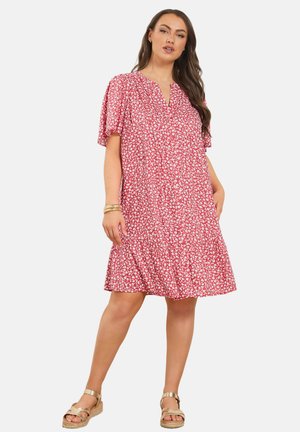 Frau trägt ein locker sitzendes rotes Kleid mit Blumenmuster und kurzen, flatternden Ärmeln, steht mit einer Hand in der Tasche und trägt goldene Sandalen und Armbänder.