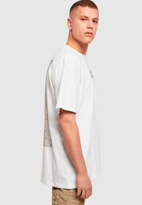 Hvid bomulds t-shirt med korte ærmer, med et puslespilsmønster på ryggen og et minimalistisk tekstdesign på forsiden.
