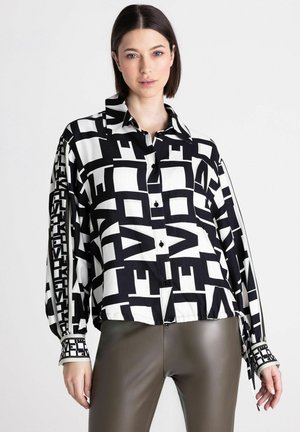 Woman with straight dark hair wears a black and white geometric patterned button-up blouse and dark brown fitted pants against a white background.