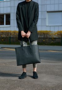 Osoba ubrana w czarną marynarkę i legginsy w kamuflaż stoi na zewnątrz, trzymając czarną torbę Ted Baker London za uchwyty.