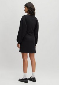 Schwarzes Sweatshirtkleid mit schmalem Taillenbereich, langen Ballonärmeln und geripptem Material. Kombiniert mit schwarzen Schuhen und weißen Socken.