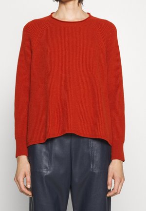 Personne portant un pull rouge côtelé à manches longues et un pantalon bleu foncé, debout devant un fond blanc uni.