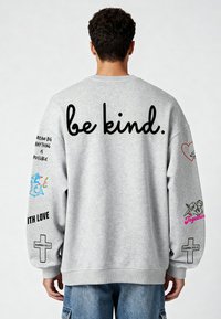 Szürke pulóver hosszú ujjakkal, amelyen nagy fekete betűkkel "legyél kedves" felirat található. A ujjain különböző grafikai nyomatok és szlogenek láthatók.