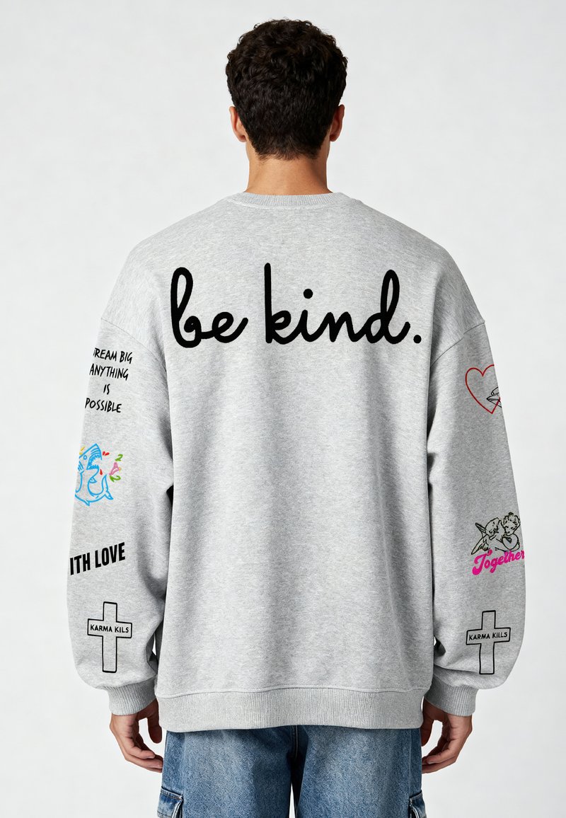 Szürke pulóver hosszú ujjakkal, amelyen nagy fekete betűkkel "legyél kedves" felirat található. A ujjain különböző grafikai nyomatok és szlogenek láthatók.