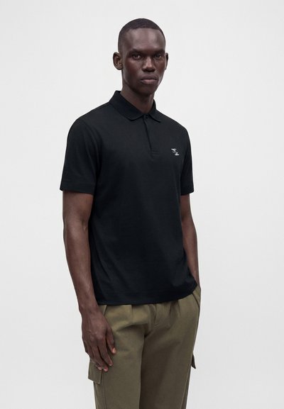 Homme portant un polo noir à manches courtes avec un petit logo blanc et un pantalon cargo vert olive, debout devant un fond uni.