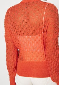 Knit sweater in bright orange with a textured open stitch pattern. Features long sleeves, a ribbed hem, and a slightly sheer back.