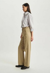 Pantalons beige à jambes larges avec une taille haute, un devant plat et des boutons accentués ; accompagnés d'une chemise à rayures légères à boutons.