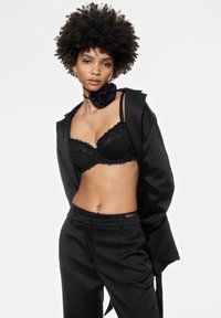 Zwarte kant beha met golvende randen, zwarte broek en een zwarte satijnen rozen chokker; model draagt een oversized zwarte blazer.