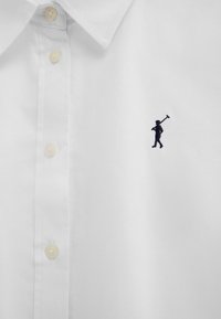 Wit overhemd met beige knopen en een klein geborduurd marineblauw logo van een persoon die een polo-knuppel vasthoudt op de borst.