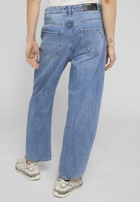 Persona vistiendo vaqueros de mezclilla azul claro holgados y zapatillas blancas y grises, de pie con la espalda hacia la cámara frente a un fondo gris.