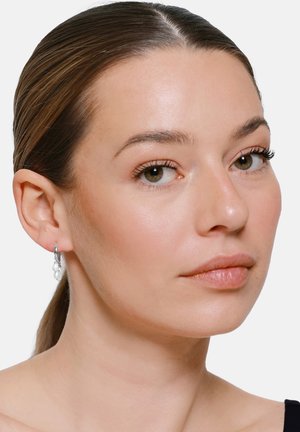 Junge Frau mit hellbraunen zurückgebundenen Haaren, trägt silberne Creolen mit kleinen weißen Perlen, schaut leicht zur Seite vor schlichtem Hintergrund.