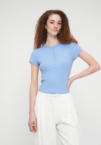 Ανοιχτό μπλε ριμπ κοντομάνικο τοπ με στρογγυλή λαιμόκοψη και διακοσμητικά κουμπιά, σε συνδυασμό με λευκό παντελόνι με κορδόνι στη μέση. Μικρή λογότυπη ένδειξη στο στήθος.