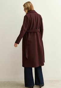Lungo cappotto bordeaux con vita cintrata e cintura, caratterizzato da un colletto e un design sartoriale. Indossato sopra pantaloni in denim scuro a zampa d'elefante. Tessuto dalla texture liscia.