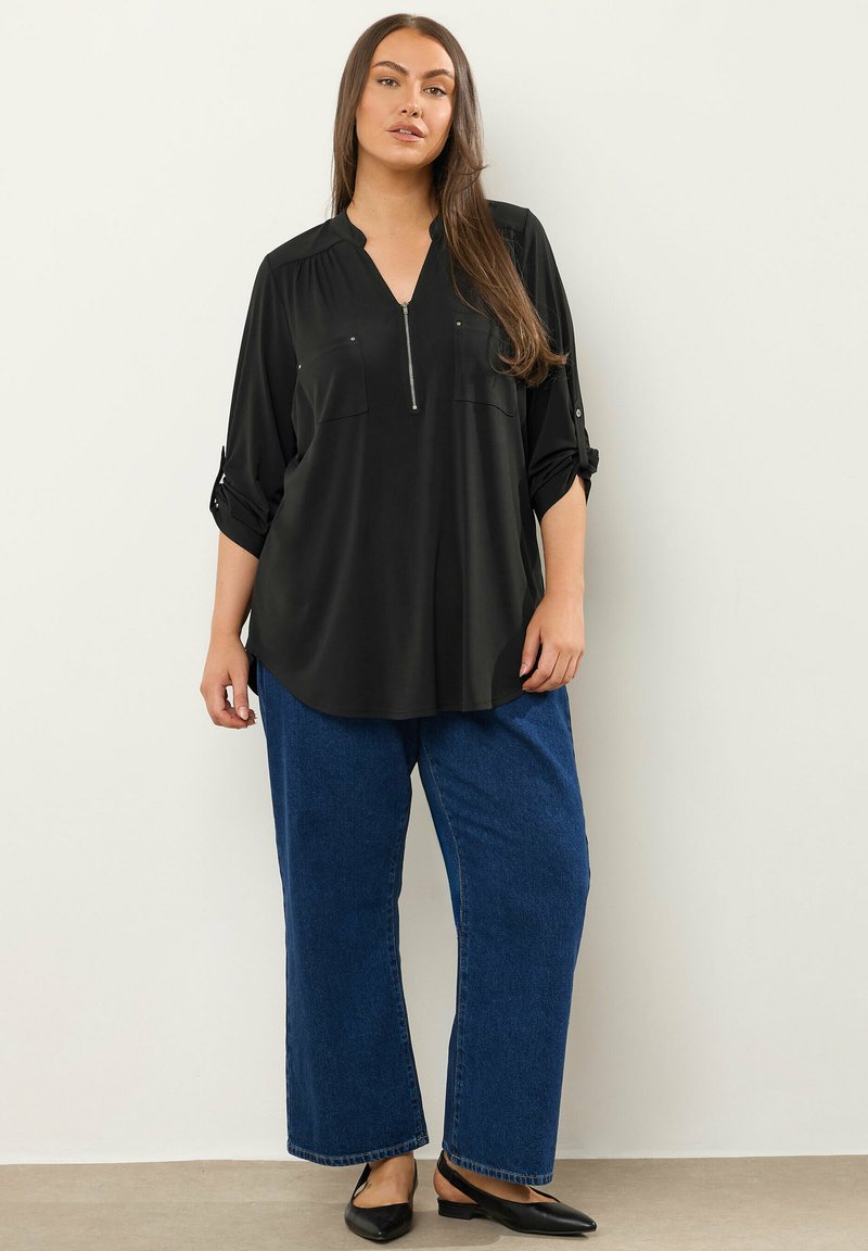 Svart blus med långa ärmar och dragkedja fram, två bröstfickor, kombinerad med vida blå denimjeans och svarta lägenheter. Slät textur.