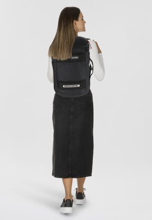 Un sac à dos noir de forme rectangulaire, avec une pièce de logo, des bretelles réglables et une surface texturée. Positionné sur le dos d'une personne.
