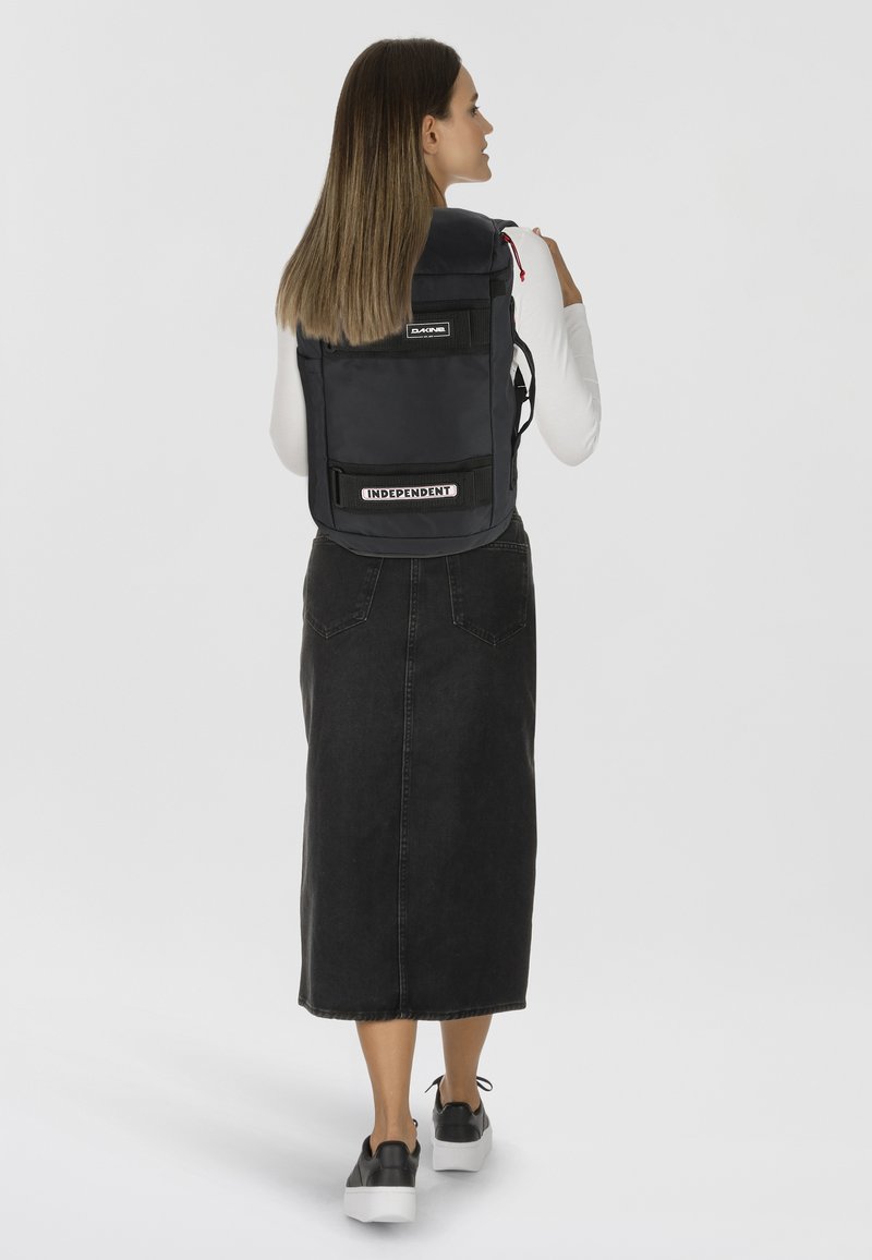 Zaino nero dalla forma rettangolare, con una patch del logo, spallacci regolabili e una superficie texture. Posizionato sulla schiena di una persona.