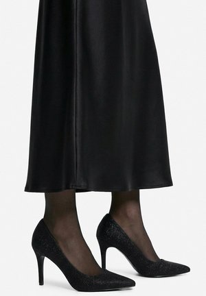 Nogi w przejrzystych czarnych rajstopach, noszące błyszczące czarne szpilki z ostrym noskiem, zestawione z czarną spódnicą midi.
