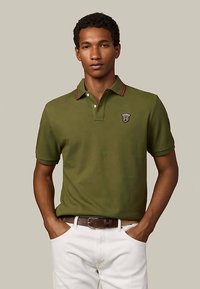 Polo vert olive avec une patte de boutonnage à deux boutons, col rouge foncé contrastant et un petit emblème sur le haut gauche. Porté avec un pantalon blanc.