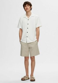 Hvit kortermet skjorte med svarte knapper, kombinert med beige shorts og lysebrune sandaler. Enkelt design, ledig passform og minimal tekstur.