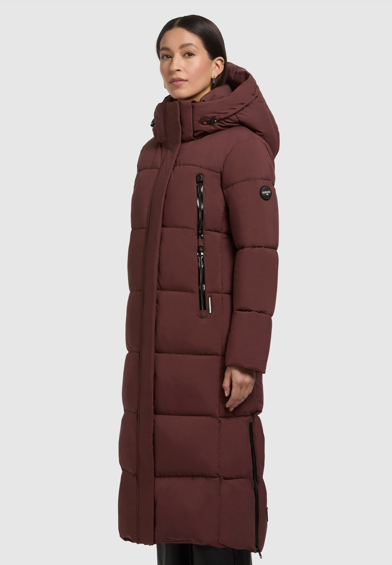 Langer burgunderfarbener Puffer-Mantel mit Kapuze, ausgestattet mit einem zipbaren Vorderreißverschluss, zwei seitlichen Taschen und gestepptem Muster zur Isolierung.