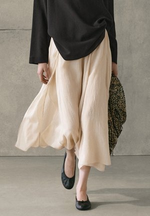 Person, die sich drinnen bewegt, trägt ein schwarzes Top, einen fließenden beigen Rock, schwarze Ballettschuhe und hält eine strukturierte Handtasche in einer Hand.