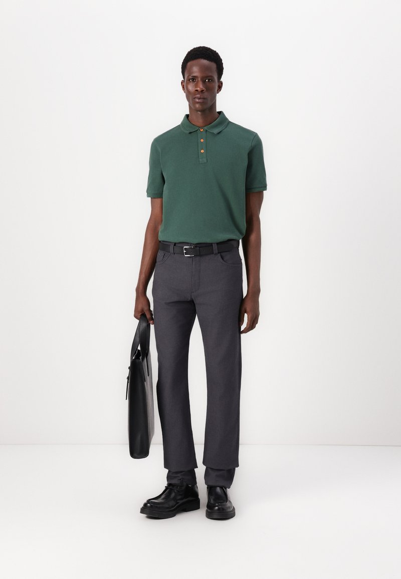 Groene polo met drie knopen, gecombineerd met grijze broek. Zwarte riem en schoenen; houdt een zwarte tas vast. Minimalistisch ontwerp, gladde textuur.