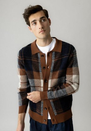 Ung mand iført en brun, marineblå og beige ternet cardigan over en hvid skjorte, kigger let opad og justerer sin ærme.