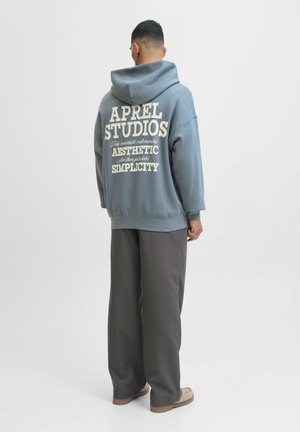 Haut à capuche bleu surdimensionné avec du texte blanc dans le dos, présentant une poche kangourou et des poignets côtelés. Assorti à un pantalon ample gris.