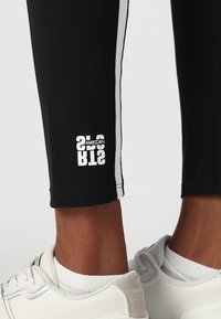 Zwarte sportleggings met witte zijstrepen, voorzien van een logo detail aan de zoom. Gladde textuur, enkel lengte en een nauwsluitend ontwerp.