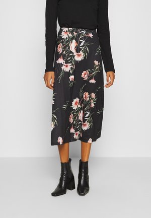 Femme portant une jupe midi noire à imprimé floral rose, associée à des bottines noires à talons et un haut noir à manches longues.