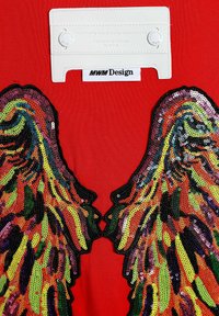 Prenda roja con alas de lentejuelas coloridas en verde, amarillo, naranja y negro, con una etiqueta blanca en el cuello que detalla la marca.