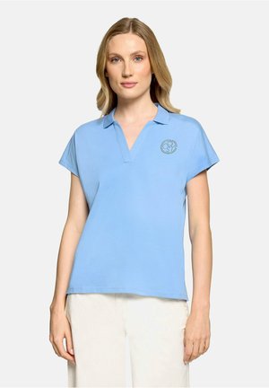 Femme aux cheveux blonds mi-longs portant une chemise bleu clair à manches courtes avec col et un pantalon blanc, debout devant un fond blanc.