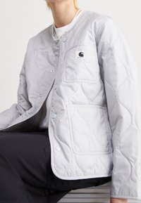 Persona vestida con una chaqueta acolchada gris claro con botones a presión, dos bolsillos delanteros y un collar de perlas, sentada contra un fondo sencillo.