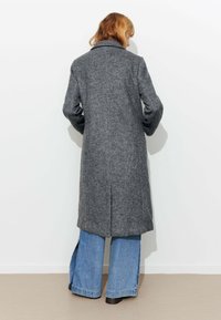 Long manteau gris en laine avec un col à revers et des fentes latérales, associé à un jean évasé bleu clair. Sur un fond blanc uni.