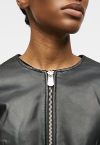 Κοντινό πλάνο του λαιμού και της γνάθου ενός ατόμου που φοράει μαύρο δερμάτινο μπουφάν με ασημένιο φερμουάρ μπροστά, μισάνοιχτο.