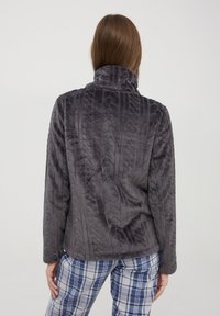 Giacca in pile grigia e strutturata con colletto alto, caratterizzata da un design a motivo. Indossata con pantaloni da pigiama a quadri.