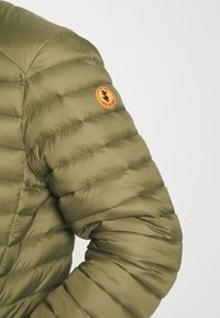 Olijfgroene pufferjack met een geborduurde inrichting. Heeft een ronde, oranje logo patch op de linkerschouder. Glad, glanzend stofoppervlak.