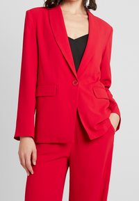 Roter Blazer aus glattem Stoff, mit Ein-Knopf-Verschluss, Revers mit Kerbe und zwei Vordertaschen. Kombiniert mit einem schwarzen Oberteil.