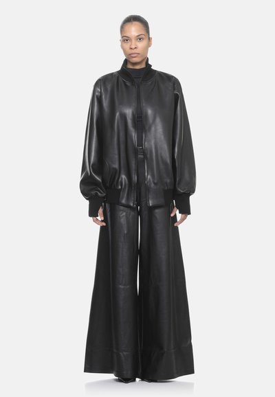 Giacca bomber in pelle nera con colletto alto e polsini a costine, abbinata a pantaloni in pelle nera a gamba larga. Texture liscia e lucida su tutta la superficie.